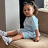 toddler, child, girl, sitting, couch, indoor, clothing, blue_outfit, sneakers, white_socks, hair_buns, hair_clips, curly_hair, face, portrait, person, furniture, modern, window, calm