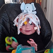 Micha joined the competition — help win amazing prizes! baby, child, high_chair, toy, blanket, elephant_plush, smiling, indoor, colorful, play, furniture, person, happy, cute, red_wheels, yellow_toy, blue_toy, hand, face, room