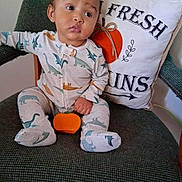 D'Andre is registered to the contest to win money with this photo: baby, child, dinosaur_pajamas, pumpkin, green_chair, cushion, pillow, text_on_pillow, indoor, cute, infant, sitting, holding_object, holiday_decor, fall_theme, portrait, person, cozy, young_child, curious