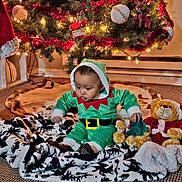 D'Andre is registered to the contest to win money with this photo: baby, child, christmas_tree, holiday, elf_costume, plush_toy, teddy_bear, blanket, carpet, lights, ornament, decorations, festive, indoor, floor, cute, green_clothing, red_and_white, toy, sitting
