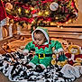 baby, child, christmas_tree, holiday, elf_costume, plush_toy, teddy_bear, blanket, carpet, lights, ornament, decorations, festive, indoor, floor, cute, green_clothing, red_and_white, toy, sitting