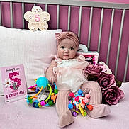 Mykha is registered to the contest to win money with this photo: baby, infant, girl, crib, pink, toys, flowers, milestone_card, headband, clothing, sitting, cute, portrait, soft_toys, pillow, blanket, indoor, child, face, decor