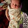 baby, sleeping, swaddled, onesie, pizza_pattern, infant, hand, adult, clothing, graphic_tee, purple_rose, skeleton_hand, closeup, indoor, cozy, soft_lighting, face, peaceful, person, portrait