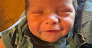 Jasper joined the competition — help win amazing prizes! baby, newborn, smiling, face, closed_eyes, camouflage, clothing, arm, hand, person, infant, skin, hair, table, paper, document, text, knitted_blanket, holding, warm_lighting