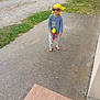 child, barefoot, yellow_bandana, red_sunglasses, toy_gun, blue_shirt, pants, outdoor, grass, concrete_path, gate, rural, dirt_road, playful, smiling, person, daylight, casual, fun, summer