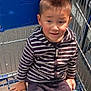 child, shopping_cart, striped_hoodie, pacifier, outdoor, parking_lot, sunlight, shadow, metal_bars, casual_clothing, young_boy, person, seat, hand, face, head, short_hair, daylight, toy, retail
