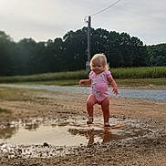 Emma is registered to the contest to win money with this photo: child, toddler, puddle, mud, splashing, pink_onesie, outdoor, dirt_path, grass, trees, nature, playing, happy, shoes, reflection, daylight, sky, field, casual, summer