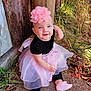 baby, child, girl, smiling, headband, pink_flower, dress, black_top, tutu_skirt, sitting, outdoor, grass, leaves, fence, wooden_post, concrete, cute, portrait, footwear, happy