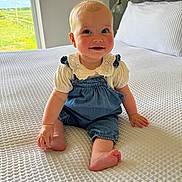 Maggie is registered to the contest to win money with this photo: baby, child, infant, denim_overalls, white_shirt, lace_collar, bed, blanket, textured_blanket, window, natural_light, indoor, smiling, happy, sitting, barefoot, head, face, skin, person