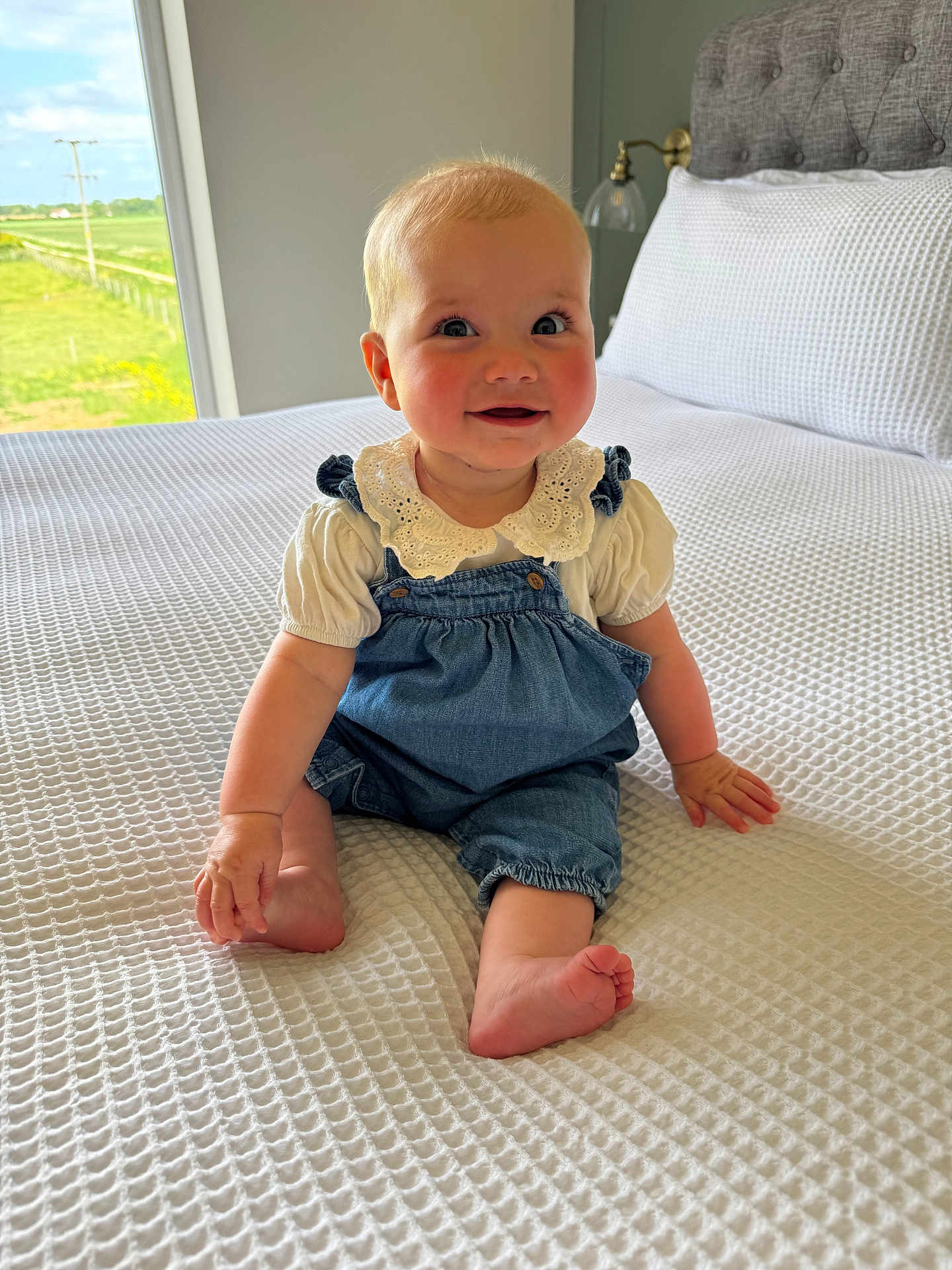 Maggie is registered to the contest to win money with this photo: baby, child, infant, denim_overalls, white_shirt, lace_collar, bed, blanket, textured_blanket, window, natural_light, indoor, smiling, happy, sitting, barefoot, head, face, skin, person