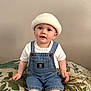 baby, beanie, casual_clothing, child, curious, cushion, cute, denim_overalls, face, hat, headwear, indoor, leaf_pattern, neutral_background, portrait, sitting, smiling, toddler, white_tshirt, young_child