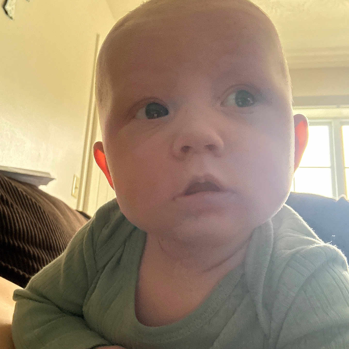 Xavien is registered to the contest to win money with this photo: baby, infant, child, face, eyes, hands, green_clothing, indoor, window, cross, light, curious, portrait, skin, head, expression, person, young, lying_down, soft_light
