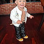 toddler, child, smiling, standing, indoor, wooden_floor, cardigan, pants, shoes, stick, brick_wall, chair, person, happy, cute, blue_eyes, blond_hair, casual_clothing, portrait, small_child