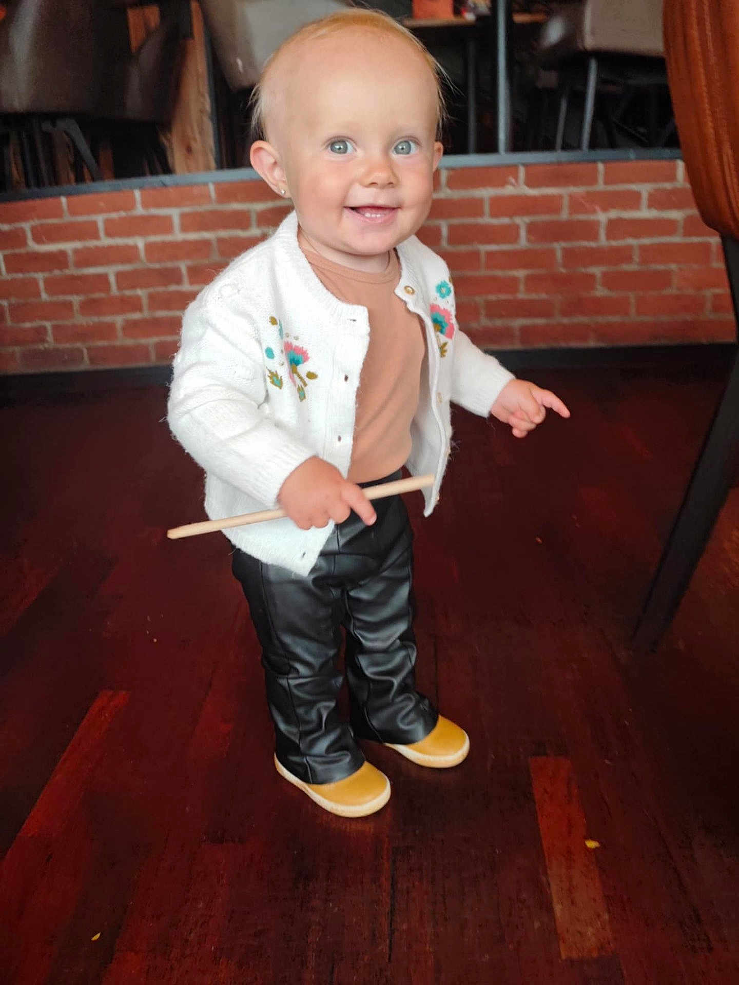 Mia participe au concours pour gagner de l'argent avec cette photo : toddler, child, smiling, standing, indoor, wooden_floor, cardigan, pants, shoes, stick, brick_wall, chair, person, happy, cute, blue_eyes, blond_hair, casual_clothing, portrait, small_child