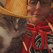 Bandit is registered to the contest to win money with this photo: cat, person, portrait, selfie, indoors, hat, straw_hat, glasses, red_clothing, hoodie, gray_cat, white_fur, sleepy, relaxed, close_up, whiskers, face, pet, companionship, humorous