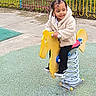 child, toddler, playground, spring_ride, yellow_horse, jacket, braided_hair, beads, outdoor, fence, concrete, rubber_flooring, black_pants, shoes, smiling, happy, play, fun, daytime, park