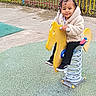 toddler, child, playground, spring_horse, yellow_toy, braided_hair, beads, jacket, black_pants, sneakers, fence, grass, outdoor, play, fun, smile, person, toy, spring, daylight