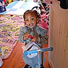 toddler, child, indoor, scooter, toy, playmat, denim_dress, purse, smiling, floor, wooden_floor, furniture, bicycle, helmet, playroom, curly_hair, black_shoes, happy, young_child, colorful