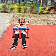Maïana participe au concours pour gagner de l'argent avec cette photo : toddler, child, swing, playground, outdoor, happy, smiling, braids, jacket, sneakers, red_surface, fence, trees, grass, park, fun, play, person, daytime, casual