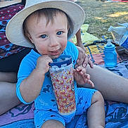 Warren a rejoint le concours — aidez-le/la à gagner de superbes lots ! toddler, child, hat, blue_outfit, cup, outdoor, grass, towel, hand, person, baby_bottle, sunhat, sitting, playful, smiling, cheeky_expression, summer, daylight, family, relaxed