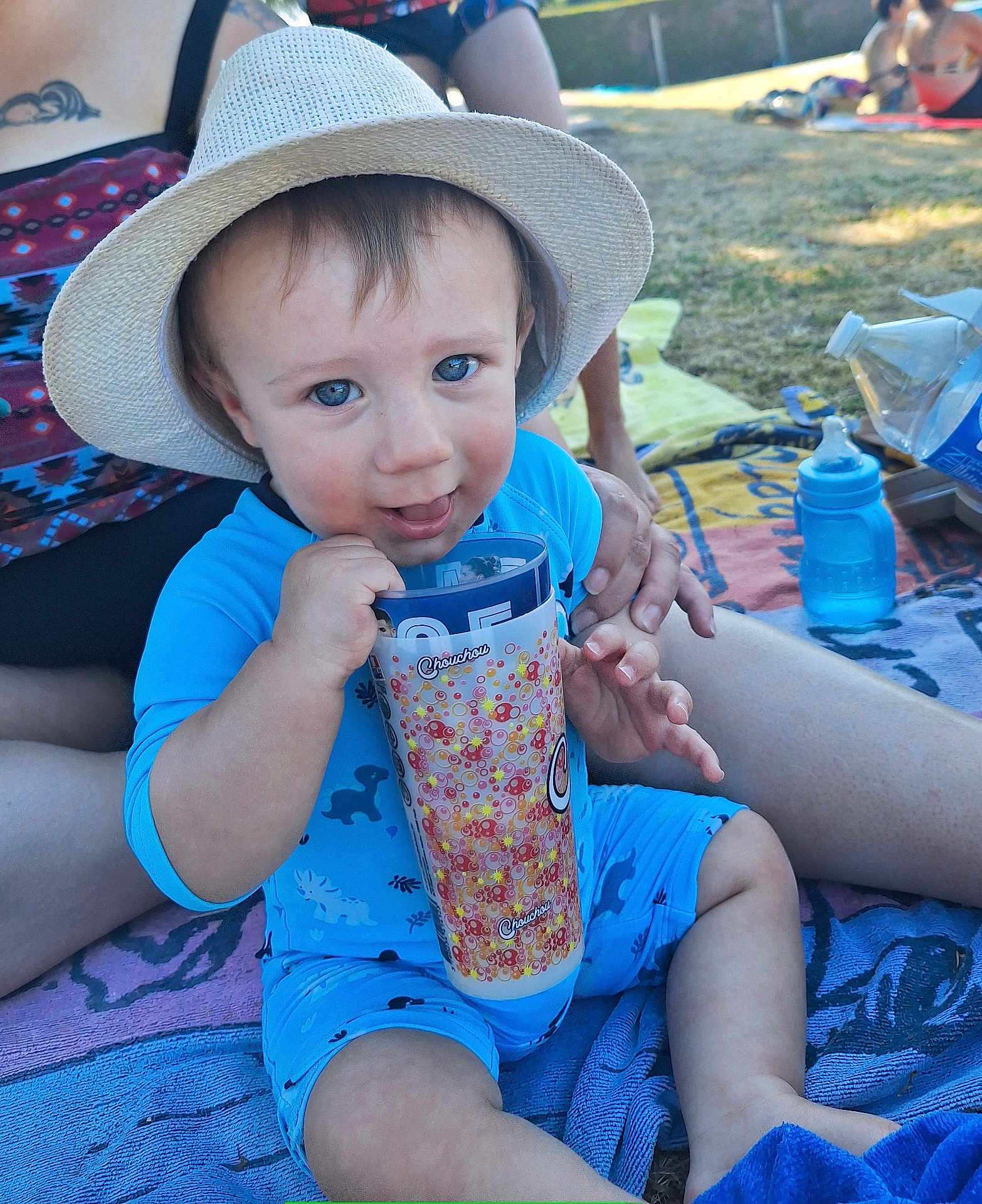 Warren a rejoint le concours — aidez-le/la à gagner de superbes lots ! toddler, child, hat, blue_outfit, cup, outdoor, grass, towel, hand, person, baby_bottle, sunhat, sitting, playful, smiling, cheeky_expression, summer, daylight, family, relaxed