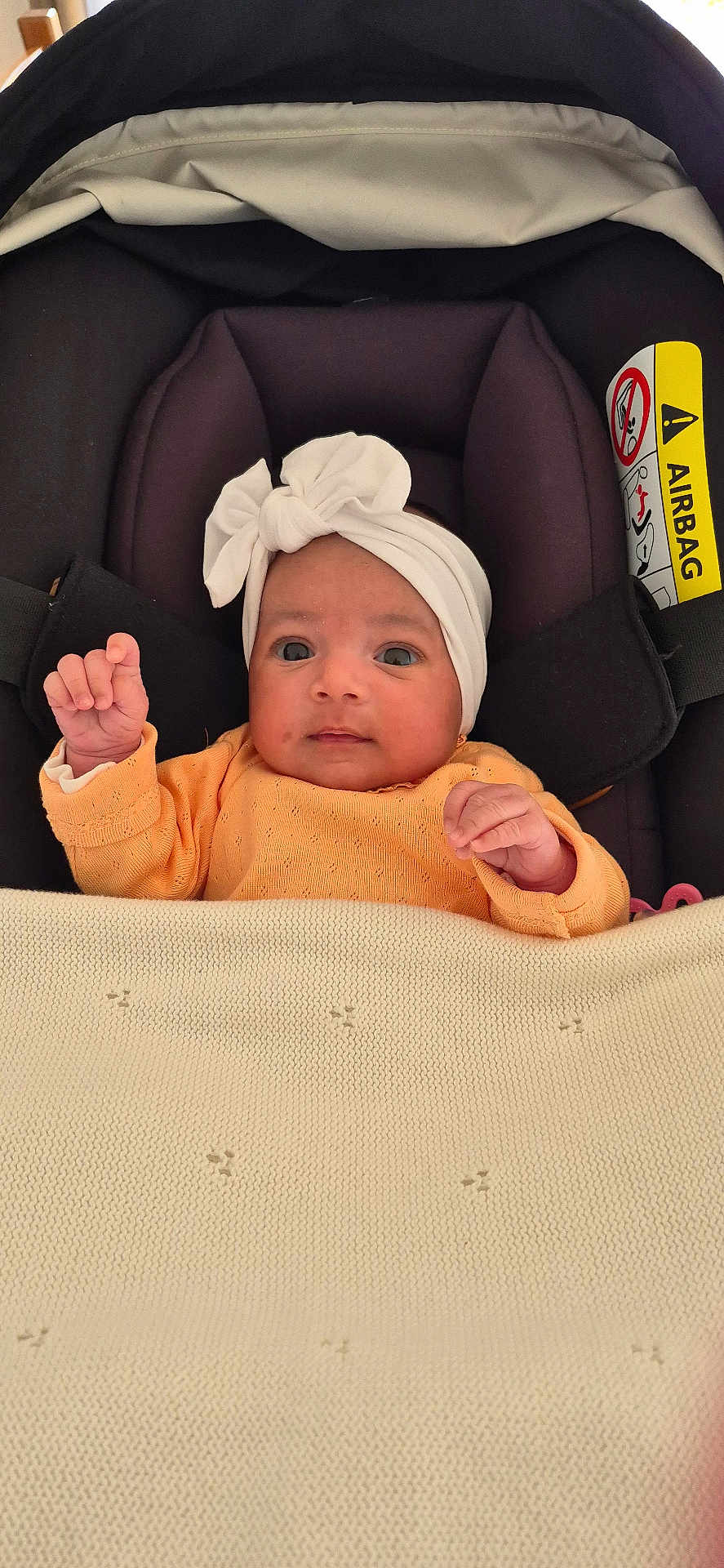 Lyianna participe au concours pour gagner de l'argent avec cette photo : baby, infant, car_seat, headband, blanket, orange_clothing, face, person, child, indoors, cute, newborn, fist, seatbelt, soft_texture, portrait, small_hands, curious, resting, cozy