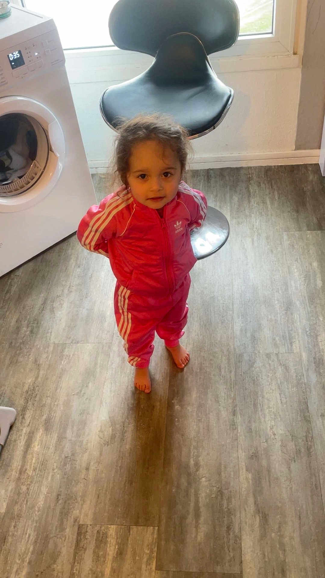 Tayana participe au concours pour gagner de l'argent avec cette photo : child, toddler, girl, pink_tracksuit, tracksuit, adidas_logo, bare_feet, washing_machine, kitchen_chair, window, laminate_floor, flooring, standing, looking_up, portrait, indoors, curly_hair, brown_hair, curious_expression, home_appliance