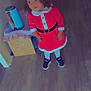 child, toddler, santa_hat, santa_costume, red_dress, curly_hair, sneakers, indoor, wooden_floor, standing, holiday, christmas, kettle, side_table, cardboard_box, drawing, portrait, cute, belt, smile