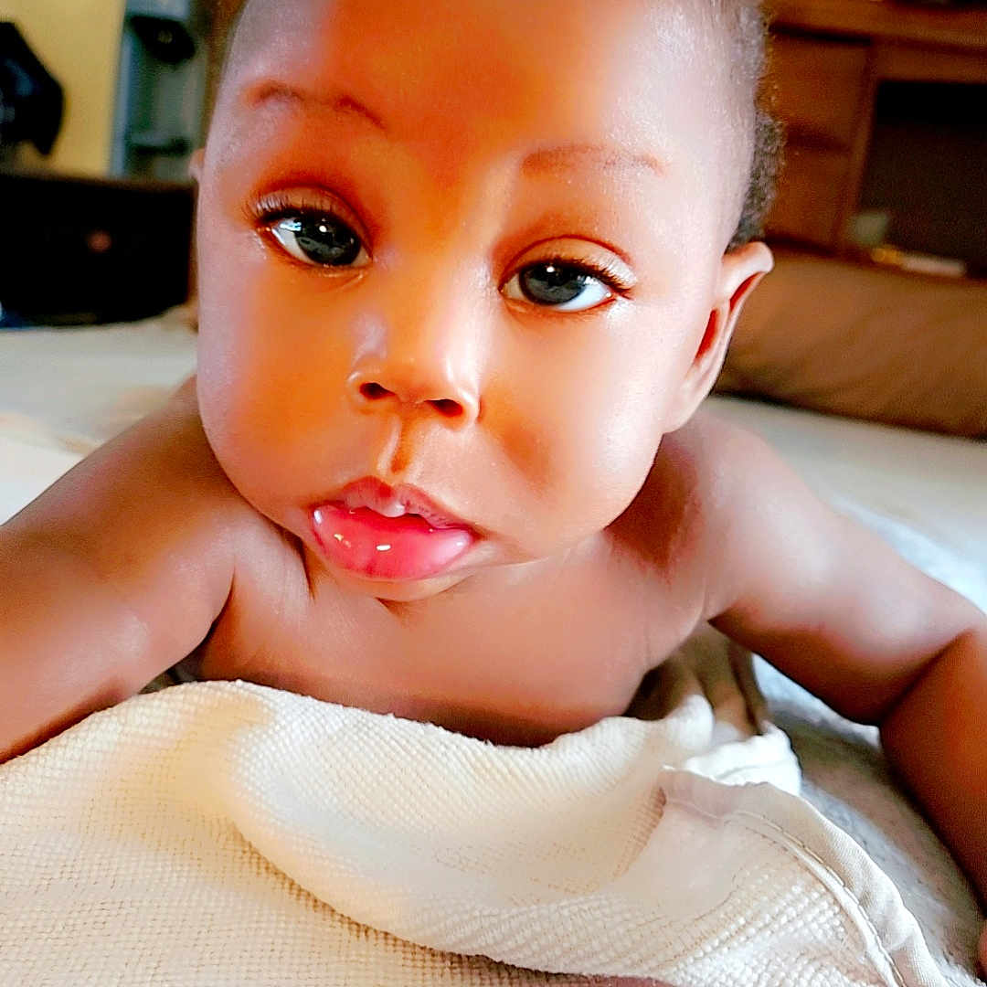 Zion is registered to the contest to win money with this photo: baby, blanket, child, closeup, curious, cute, drool, expression, eyes, face, furniture, homedecor, indoors, infant, lying_down, portrait, skin, soft_texture, topknot, warm_lighting