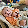 baby, baby_seat, blanket, child, cozy, cushion, doll, face, furniture, hand, happy, home, indoor, infant, orange_clothing, person, relaxed, smiling, soft_toy, toy