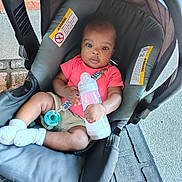 Shane is registered to the contest to win money with this photo: baby, bottle, car_seat, child, clothing, coral_shirt, floor, footwear, infant, pacifier, person, plastic_bottle, socks, seat, seatbelt, sidewalk, sitting, skin, toy, young_child