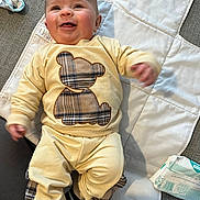 Blake is registered to the contest to win money with this photo: baby, infant, smiling, yellow_pajamas, plaid, teddy_bear, changing_mat, diaper_pack, happy, cute, child, person, clothing, indoor, floor, baby_care, lying_down, small_hands, footwear, joyful
