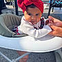 Cataleya participe au concours pour gagner de l'argent avec cette photo : baby, child, headwrap, red_headwrap, white_sweater, baby_walker, outdoor, sunlight, hands, person, curious_expression, footwear, black_shoes, plaid_skirt, asphalt, chair, table, vehicle, support, infant