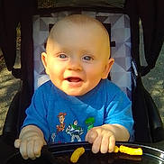 Sterling is registered to the contest to win money with this photo: baby, blue_eyes, cheese_puffs, child, clothing, cute, face, hands, happy, infant, outdoor, person, portrait, seat, smiling, snack, stroller, sunlight, toy_story_shirt, tray