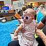 baby, child, indoor, play_area, sunglasses, pink_clothing, striped_clothing, adult_hand, pirate_ship, blue_carpet, hospital_sign, people, woman, tattoo, headband, toy, shopping_mall, casual_clothing, cute, childrens_hospital