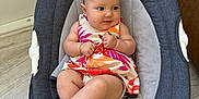 Sophia is registered to the contest to win money with this photo: baby, infant, baby_rocker, dress, colorful_clothing, indoor, floor_tiles, baby_seat, child, cute, plush_fabric, curly_hair, barefoot, sitting, bracelet, earrings, modern_furniture, neutral_colors, looking_away, soft_lighting