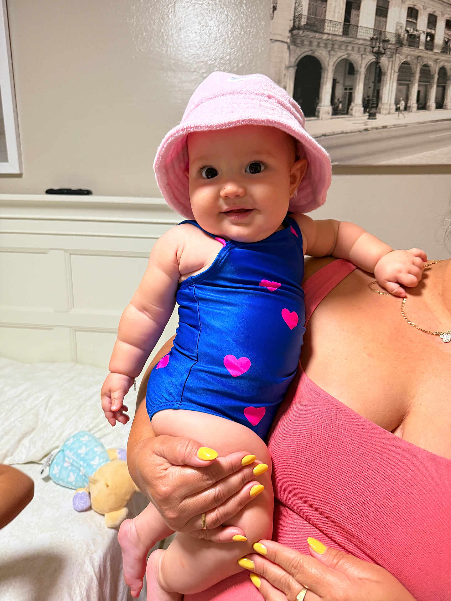 Sophia joined the competition — help win amazing prizes! baby, child, pink_hat, blue_swimsuit, hearts, adult_hand, yellow_nails, indoor, bed, plush_toy, portrait, smiling, cute, holding, person, skin, fingers, jewelry, nail_polish, room