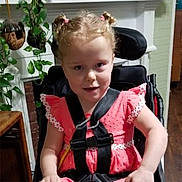 Aurora is registered to the contest to win money with this photo: child, girl, wheelchair, indoor, pink_dress, blonde_hair, hair_buns, straps, plant, wooden_furniture, smiling, seated, young, face, person, home, decor, bright_clothing, cute, portrait