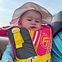 baby, child, life_jacket, hat, sunhat, safety, outdoor, water_sport, clothing, person, infant, expression, hand, seat, blue_sleeves, yellow_strap, pink, yellow, gray, portrait
