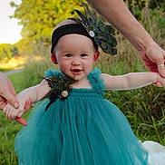 Vivian is registered to the contest to win money with this photo: baby, child, dress, teal_dress, peacock_feathers, headband, outdoor, grass, nature, adult_hands, holding_hands, smiling, portrait, infant, happy, person, greenery, field, cute, sunlight