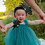 baby, child, dress, teal_dress, peacock_feathers, headband, outdoor, grass, nature, adult_hands, holding_hands, smiling, portrait, infant, happy, person, greenery, field, cute, sunlight