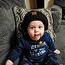 Khaled is registered to the contest to win money with this photo: baby, child, beanie, sweatshirt, text_clothing, couch, pillow, gray, pattern, indoor, smiling, cute, person, sitting, young_child, face, eyes, hand, clothing, portrait