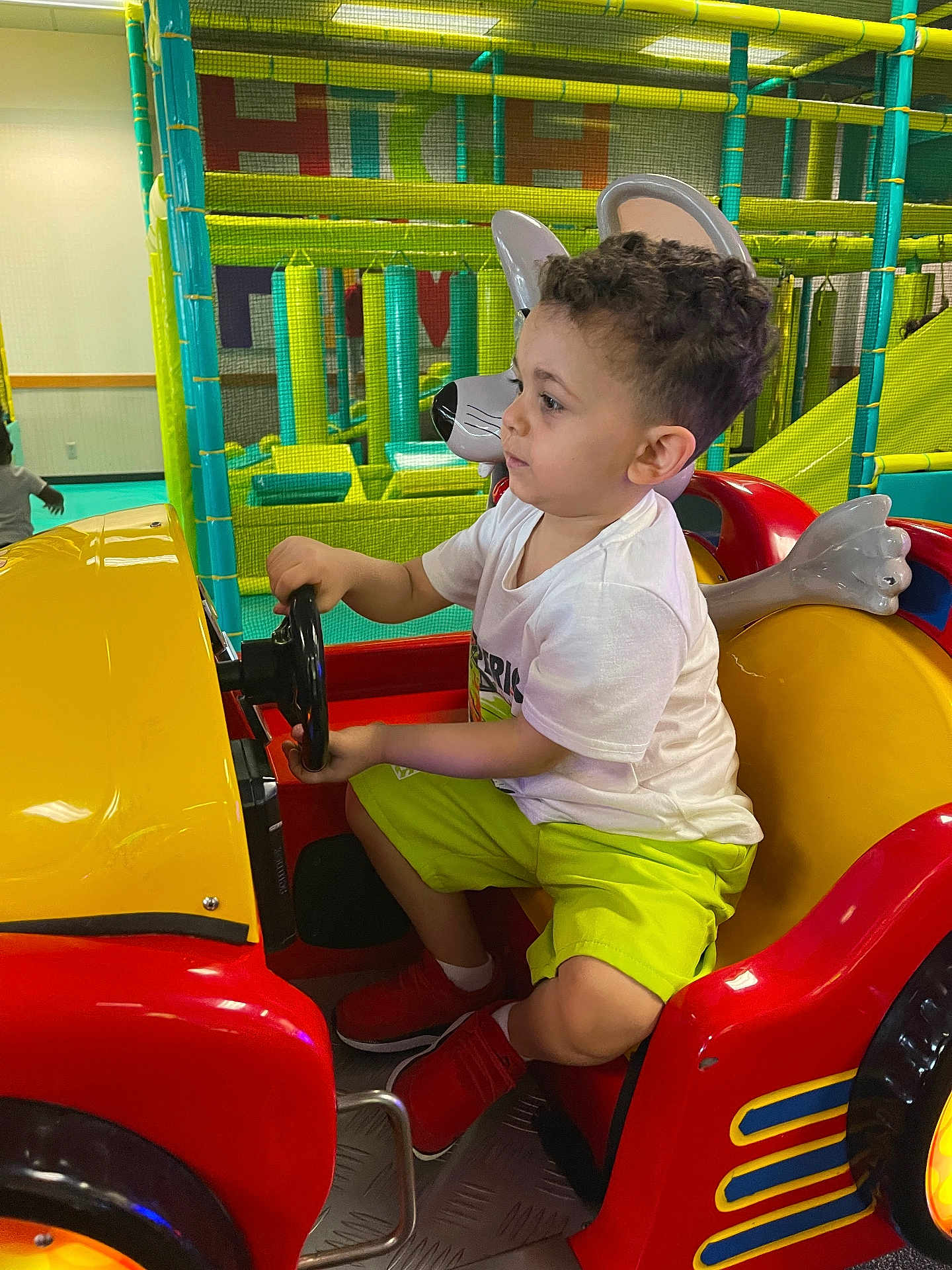 Khaled joined the competition — help win amazing prizes! child, kid, indoor_play_area, kiddie_ride, toy_car, steering_wheel, playground, colorful, yellow, red, green, shorts, tshirt, curly_hair, footwear, playing, fun, entertainment, boy, side_view
