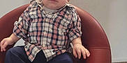 Khaled is registered to the contest to win money with this photo: baby, child, plaid_shirt, red_chair, sitting, indoor, carpet, curious, person, casual_clothing, short_hair, infant, neutral_expression, modern_furniture, footwear_bare, small_hands, cute, portrait, young, home
