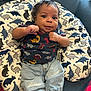 baby, infant, dinosaur_print, clothing, jeans, socks, pillow, couch, curly_hair, cute, indoor, portrait, child, furniture, pattern, textile, smiling, person, casual_wear, seated