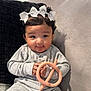 Zipporah is registered to the contest to win money with this photo: baby, child, bow, hair_accessory, gray_outfit, teething_toy, couch, pillow, indoor, cute, smiling, person, infant, sitting, hands, face, black_pillow, cozy, playful, adorable