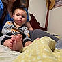baby, child, adult, bed, blanket, bottle, foot, pajamas, person, furniture, headboard, hair, face, hand, sitting, indoor, clothing, toy, yellow_blanket, blue_shirt