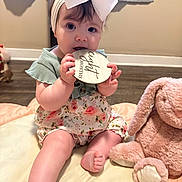 Ivee is registered to the contest to win money with this photo: baby, child, infant, headband, bow, plush_toy, pink, floral_dress, sitting, chewing, wooden_plaque, eight_months, soft_mat, cute, indoors, floor, toy, person, young_child, playtime