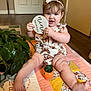 baby, child, nine_months, quilt, headband, butterfly_pattern, plant, potted_plant, indoor, floor, wooden_sign, happy, smiling, sitting, legs, feet, hands, curly_hair, cute, infant