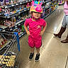 child, helmet, pink_clothing, toy_store, shopping_cart, floor, sneakers, shorts, tshirt, person, aisle, shelf, toys, shopping, helmet_unicorn, standing, retail, supermarket, casual, cute
